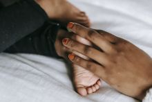 Unrecognizable African American mother touching barefoot foot of faceless infant baby while lying on white blanket in bedroom on bed